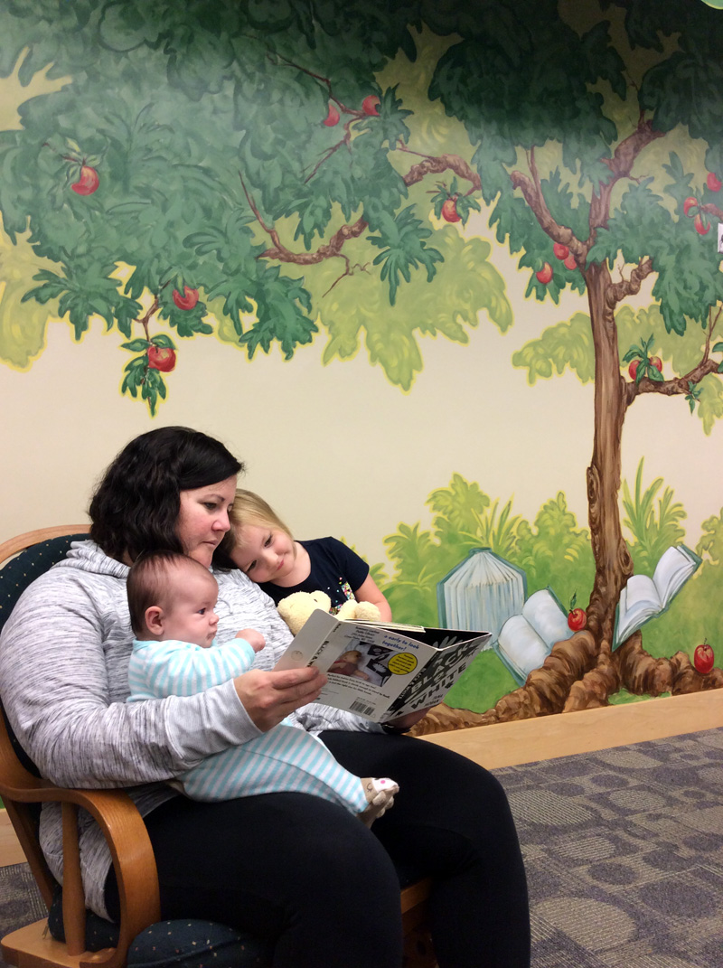 Mom reads to young daughter and baby in a rocking chair in front of apple tree mural.