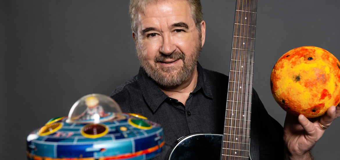 Singer/Storyteller Randy Peterson holding guitar, planet and flying saucer