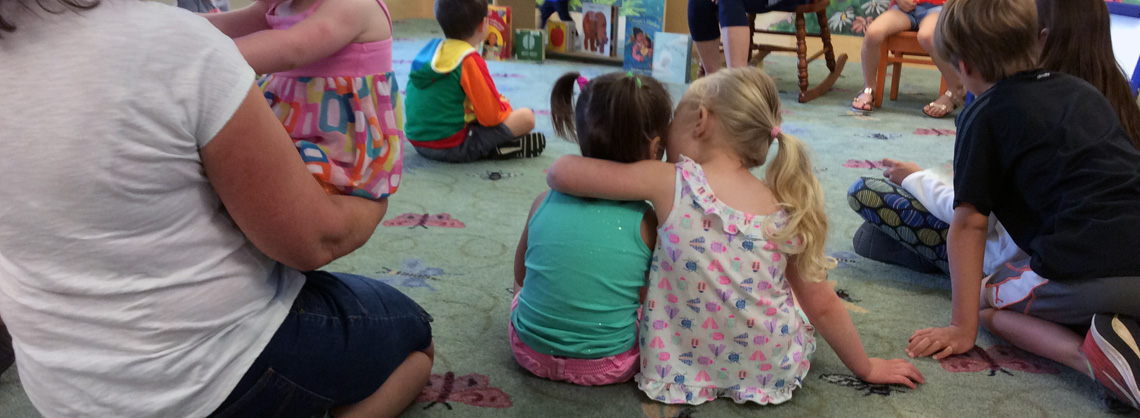 two little girls, one with her arm around the other's shoulders, listen to stories